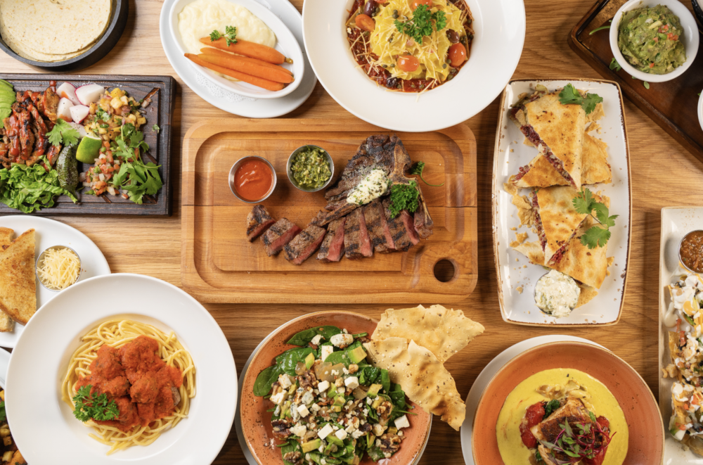 This image shows a sumptuous spread of various dishes from Rascal Modern American Diner & Bar in Sedona, Arizona. The table is covered with an assortment of beautifully presented meals including grilled steak with a side of chimichurri sauce, a plate of spaghetti topped with meatballs, nachos garnished with shredded cheese and tomatoes, seared fish atop a creamy yellow sauce, a colorful garden salad with nuts and cheese, quesadillas cut into triangles, a bowl of guacamole beside some chips, and mashed potatoes alongside glazed carrots. The presentation is vibrant and inviting, showcasing a diverse menu that caters to different tastes.